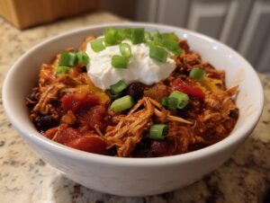 A close-up of a white bowl filled with shredded Turkey chili, topped with sour cream, cheese, and green onions.