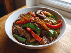 A close-up of a white bowl filled with glossy Amazing 30 Minute pepper steak featuring beef strips and vibrant red and green bell peppers.