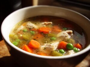 Close-up of a white bowl filled with amazing chicken vegetable soup, showing broth, carrots, peas, and shredded chicken.