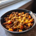 A close-up of a savory sweet potato skillet dish featuring ground meat and melted cheddar cheese in a cast iron pan.