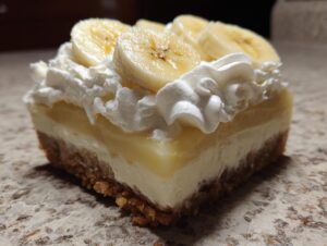 A close-up of a square slice of banana cream cheesecake with graham cracker crust, cream cheese layer, banana pudding, and whipped cream topping with banana slices.