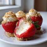 Four cheesecake deviled strawberries on a white plate, one cut open revealing creamy filling and graham cracker crust base.