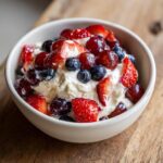 A bowl filled with creamy cheesecake fruit salad topped with fresh strawberries, blueberries, and red grapes.