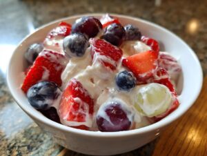 A close-up view of a white bowl filled with creamy cheesecake fruit salad featuring strawberries, blueberries, and grapes.