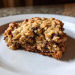 A close-up, tasty shot of a thick, chewy chocolate chip oatmeal cookie bar resting on a white plate.