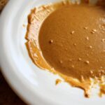 A close-up, overhead shot of thick, creamy peanut sauce with visible specks, swirled in a white bowl.