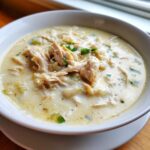 A close-up of a hearty bowl of creamy, white cream cheese chicken chili topped with shredded chicken and green onions.