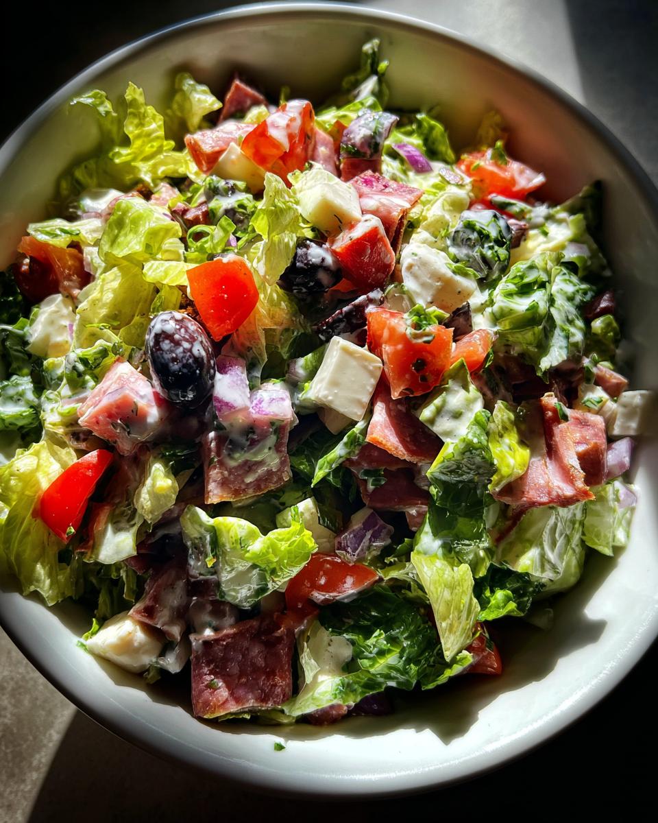 A vibrant bowl of grinder salad featuring lettuce, salami, cheese cubes, tomatoes, and olives tossed in a creamy dressing.