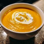 Close-up of a bowl of vibrant orange creamy pumpkin soup, topped with a swirl of cream and cracked pepper.