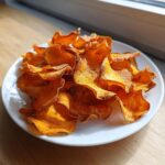 A pile of crispy, orange sweet potato chips seasoned with spices, served on a white plate.