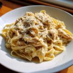 A close-up of creamy fettuccine pasta mixed with shredded chicken and topped generously with grated parmesan cheese.