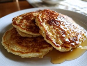 Close-up of a stack of three golden brown almond flour pancakes drizzled generously with maple syrup.