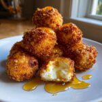 A stack of crispy, golden brown fried goat cheese balls drizzled with honey, one is cut open showing the soft interior.