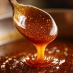 Close-up of thick, glossy homemade caramel sauce dripping from a wooden spoon back into the pot.