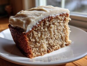 Close-up of a moist slice of applesauce cake topped with thick, spiced cream cheese frosting on a white plate.