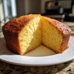 A golden brown olive oil cake, cut in half to show its moist, yellow crumb, resting on a white plate.
