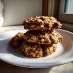 Three textured oatmeal breakfast cookie stacked on a white plate, highlighted by strong sunlight.