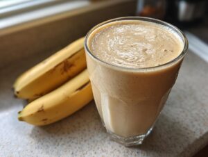 A tall glass filled with a creamy peanut butter banana smoothie next to two whole bananas.