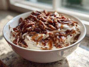 A white bowl filled with creamy pecan pie dip topped generously with glazed pecans and caramel sauce.