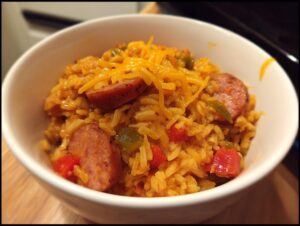 Close-up of a white bowl filled with flavorful sausage and rice mixed with peppers and topped with melted shredded cheddar cheese.
