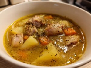 Close-up of a white bowl filled with savory sauerkraut soup featuring chunks of meat, potatoes, and carrots, seasoned with herbs.