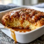 A close-up of a warm snickerdoodle cobbler served in a white dish, topped generously with gooey caramel sauce.