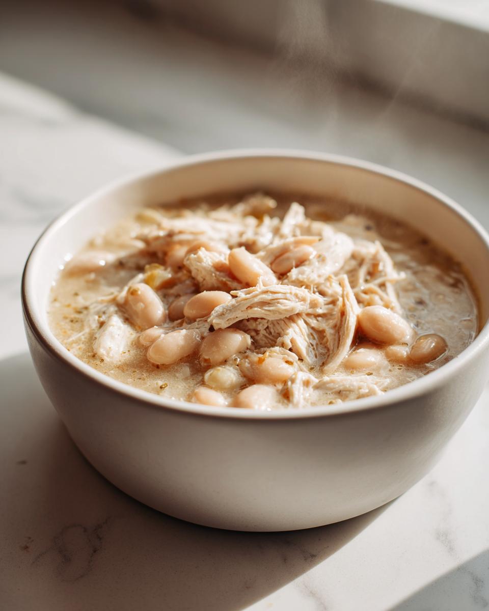 Close-up of a steaming white bowl filled with white bean chicken chili, featuring shredded chicken and white beans in a creamy broth.
