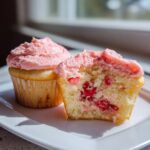 Two strawberry cupcakes on a white plate, one cut in half showing the moist cake filled with strawberry pieces.