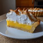 A perfect slice of lemon meringue pie showing bright yellow filling, flaky crust, and tall, torched meringue peaks.