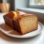 A loaf of moist sugar free banana bread, with one thick slice cut and displayed on a white plate.