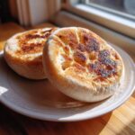 Two freshly cooked homemade english muffins with characteristic nooks and crannies, lightly browned on a plate.