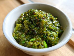 Close-up of a thick, vibrant green tomato salsa with visible herbs and chili flakes in a small white bowl.