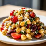 A vibrant mound of mediterranean chickpea salad featuring chickpeas, halved cherry tomatoes, Kalamata olives, and crumbled feta cheese.
