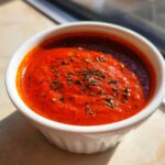 Close-up of rich, red pizza sauce topped with dried herbs in a small white bowl.