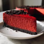 A close-up shot of a rich, vibrant red velvet cheesecake slice on a white plate, topped with dark chocolate crumbs.
