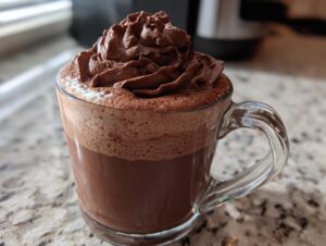 A clear glass mug filled with rich, frothy whipped hot chocolate topped with a swirl of chocolate whipped cream.