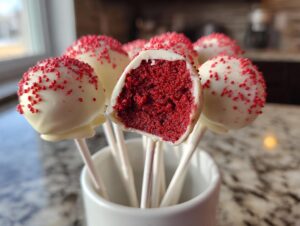 A cluster of white chocolate-dipped red velvet cake pops decorated with red sprinkles, one cut open showing the vibrant red interior.
