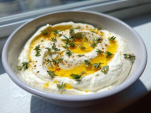 A bright white bowl filled with creamy ricotta dip, drizzled with olive oil and fresh thyme.