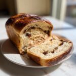 A golden-brown loaf of amazing 1-step raisin bread, partially sliced on a white plate.