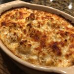 Close-up of a bubbling, baked hot crab dip with a golden-brown, cheesy crust in an oval ceramic dish.