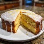 A moist, two-layer lemon cake recipe with thick white glaze and lemon zest, served on a white plate.