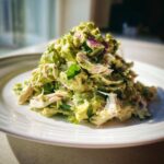 A generous mound of creamy avocado chicken salad featuring shredded chicken, chunks of avocado, and red onion on a white plate.