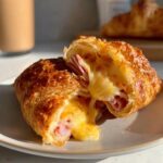 Close-up of a baked ham and cheese croissant cut in half, showing melted cheese stretching out.