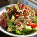 A bowl of creamy blt chicken salad served over lettuce, topped with avocado chunks and crispy bacon bits.
