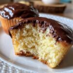 Close-up of a boston cream pie cupcake with a bite taken out, showing the fluffy yellow cake and rich chocolate glaze.