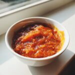Close-up of a small white bowl filled with thick, chunky orange chili cheese dip, illuminated by sunlight.