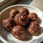 A small white bowl filled with several rich, dark chocolate protein balls studded with visible oats.
