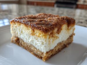 A square slice of churro cheesecake bar featuring a graham cracker crust, creamy white filling, and cinnamon sugar topping.