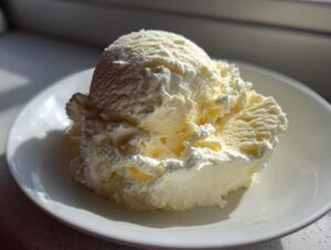 Two scoops of pale yellow, creamy cottage cheese ice cream served on a white dish, highlighted by sunlight.
