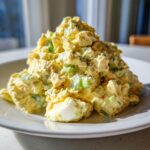 A close-up, appetizing mound of creamy egg salad recipe mixed with chopped green onions on a white plate.